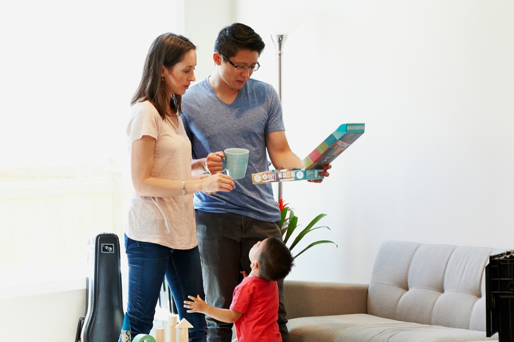Two adults talking at home while a toddler plays with blocks