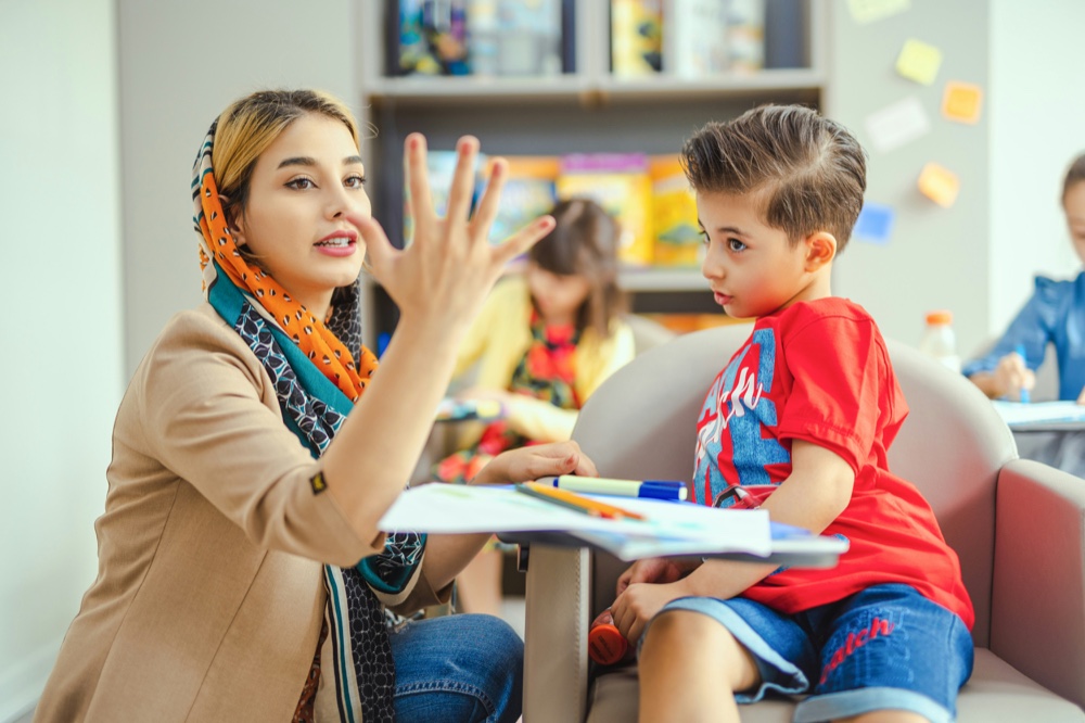 An adult and a child talking across a table in a busy classroom